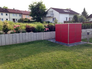 . The image depicts a neat suburban backyard scene. In the foreground, there is a grassy lawn with a small, modern, red utility or storage structure, possibly a shed. Behind it, there is a low, gray stone retaining wall. Beyond the wall, you can see a row of houses with well-maintained gardens and blooming flowers across green foliage. The houses have tiled roofs and are situated in a residential area. There is also an outdoor lamp post visible to the right, indicating it might be a backyard or garden area.