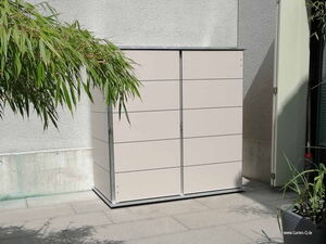 . Ein weißer Gartenschrank steht draußen auf einer Terrasse neben Pflanzen und einer Wand.