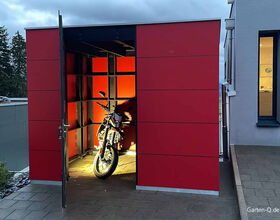 . Rotes Gartenhaus mit offenem Tor, darin ein beleuchtetes Motorrad.