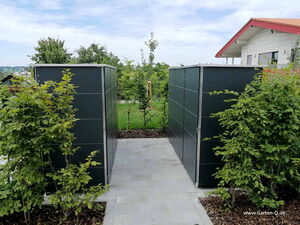. Ein moderner Gartenschuppen aus Metallplatten, umgeben von Bäumen und einem gepflasterten Weg.