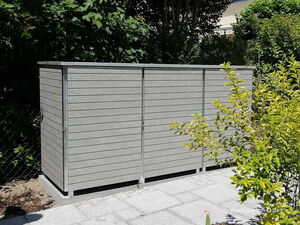 . Ein grauer Gartenschrank mit Lamellen steht auf einem gepflasterten Platz, umgeben von Bäumen und Sträuchern.