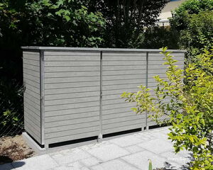 . The image shows a rectangular storage unit or shed with horizontal wooden slats, placed on a paved surface. It is situated next to some greenery, including bushes and trees. The design looks modern and is likely used for outdoor storage.