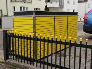 . The image shows a bright yellow storage unit with horizontal slats. It's situated outdoors, next to a fence with black vertical bars and yellow caps on top. The background includes urban elements like a building and part of a parked blue car.
