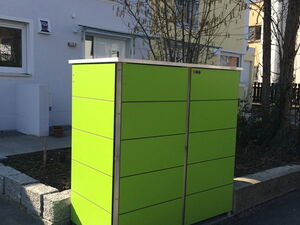 . The image shows a bright green outdoor enclosure, likely a bicycle storage box, situated on a sidewalk near a residential area. It's placed parallel to the road, with a building in the background and some landscaping around it. The enclosure seems to be made of metal panels and has a flat top.