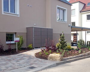. The image shows the exterior of a modern residential building with neatly landscaped surroundings. There is a pathway leading to the entrance, bordered by garden beds with various plants and shrubs. A small sign is visible near the plants. The facade has several windows, and the overall appearance is clean and well-maintained.