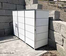 The image shows a modern, sleek outdoor storage unit placed against a backdrop of large stone blocks. The unit has a minimalist design with clean, white panels and seems to be part of a tidy, landscaped outdoor area.
