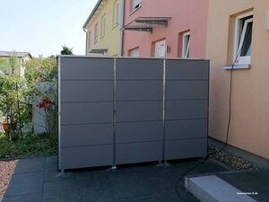 . The image shows a modern outdoor storage unit or garden shed with a sleek, gray panel design. It's placed on a patio near the wall of a house with colorful exterior walls. There are some plants visible next to the storage unit, and a cable connected to a nearby window. The ground is covered with a mix of stone paving and gravel.