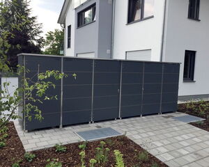 . The image shows a modern structure with a sleek, dark exterior. It appears to be a series of storage units, possibly for trash bins or outdoor equipment, next to a building. The structure is located on a paved area surrounded by a garden with plants. The building next to it is a contemporary-style house.