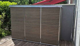 Ein Gartenbereich mit Holzplattform, Abfalleinhausung aus Holz und Metall, und roter Markise.