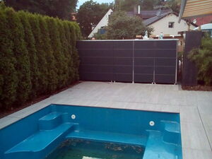 . The image shows an empty swimming pool with a modern design, featuring built-in steps. The surrounding area is a tiled patio with trees and shrubs along one side, providing some greenery. In the background, there are residential houses partially visible, and an extended awning is at the top-right corner, suggesting a partially covered area. The setting appears to be a backyard or a private outdoor space. 