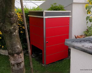. The image shows a red garden storage unit with a sleek, modern design situated outdoors. It is placed near a tree and surrounded by yellow flowers and greenery. There are some wooden sticks leaning against the tree, and the structure seems to be on a lawn area.