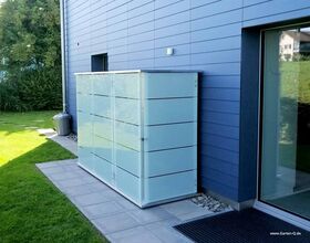 . The image shows a sleek outdoor storage unit with a contemporary design, positioned next to a blue-paneled house. It's placed on a tiled area adjacent to a glass door, with a modern facade matching the style of the building. The area around it is grassy and landscaped.