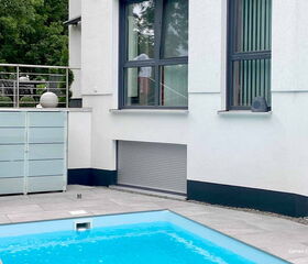Moderner Garten mit Terrasse, Pool und Glasgeländer neben einem weißen Gebäude.