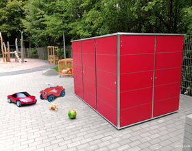 Roter Schuppen, Spielzeugautos, Teddy und Ball auf Pflastersteinen vor einem Spielplatz mit Bäumen im Hintergrund.