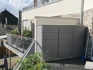 . Ein Balkon mit Pflanzkästen, Geländer und einer grauen Box. Gebäude und Himmel im Hintergrund.