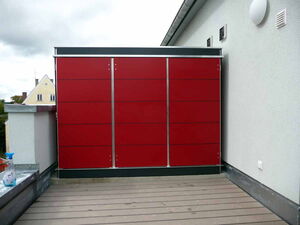 . The image shows a modern, red outdoor cabinet-like structure on a balcony or terrace. It's set against a light-colored wall, and there's a wooden deck in the foreground. A building with a gabled roof is visible in the background.