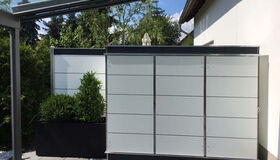 . Terrasse mit Sichtschutzwänden aus Glas und Pflanzenkübel, überdacht mit Glasdach, umgeben von Bäumen.