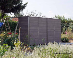 . The image shows a garden scene featuring a modern, rectangular garden shed with sleek, dark wood paneling. There's a play area in the background with a wooden swing set containing a slide and swings. The area is surrounded by various plants and bushes, and there's a chain-link fence in the distance. The website URL "www.Garten-Q.de" is visible in the bottom right corner.