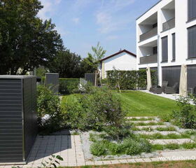 . The image shows a modern garden area adjacent to a building. The scene includes:

1. A lawn with neatly trimmed grass.
2. Several small shrubs and plants.
3. A paved walkway.
4. Two metal garden storage units.
5. A multi-story building with balconies and large windows.
6. Patio furniture arranged on a terrace.
7. Trees and hedges in the background.

The overall setting appears to be well-maintained and landscaped, suggesting a residential or apartment area.